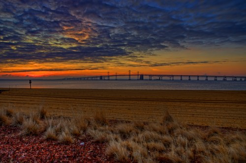 Sunrise over a bridge spanning a calm body of water, with sandy beach and grass in the foreground under a colorful sky.
