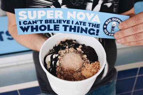 A person holds a bowl of ice cream topped with various ingredients and a sign that reads "SUPER NOVA I can't believe I ate the whole thing!"
