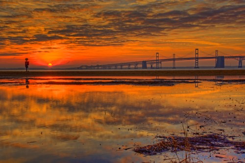 A vibrant sunset reflects on water, with a bridge silhouetted against the colorful sky and clouds.