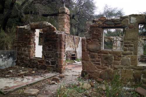 Ruins of a stone building surrounded by overgrown vegetation and trees, with partially collapsed walls and a weathered appearance.