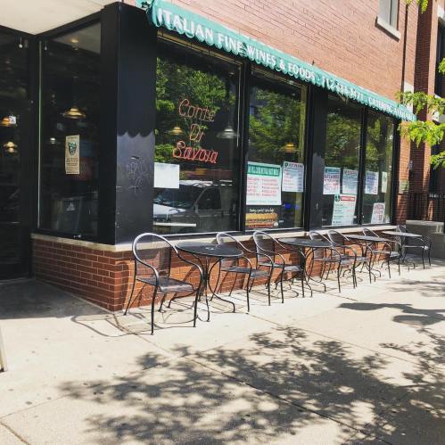 A storefront with large windows displaying menus and outdoor seating in front on a sunny day.