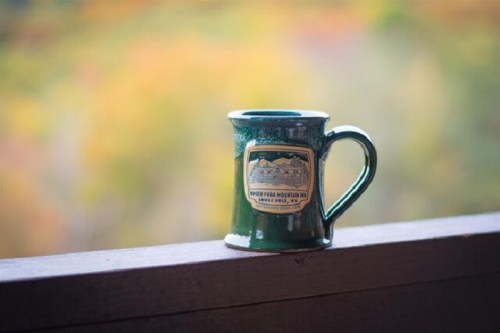 A green ceramic mug with a label sits on a wooden railing, with a blurred colorful background of trees.