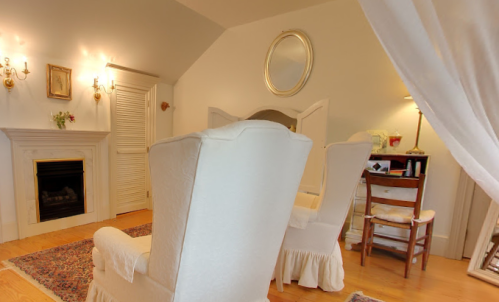 Cozy room with two white chairs, a fireplace, a mirror, and a small desk, featuring soft lighting and warm decor.