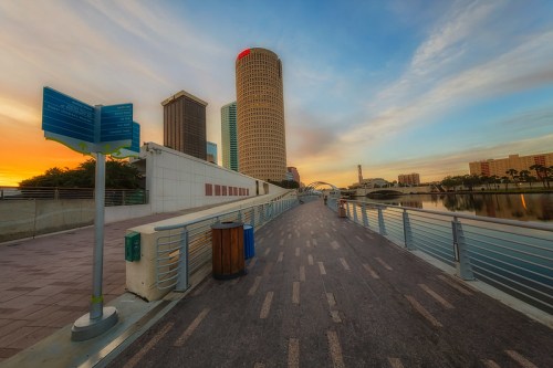 A scenic waterfront walkway with buildings and a sunset in the background, featuring a clear sky and calm water.