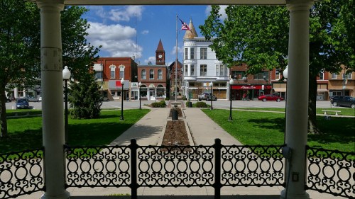 A view of a town square with a clock tower, American flag, and surrounding buildings, framed by a decorative gazebo.