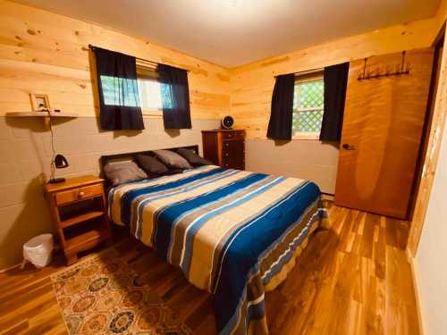 Cozy bedroom with wooden walls, a striped bedspread, nightstand, dresser, and two windows with dark curtains.