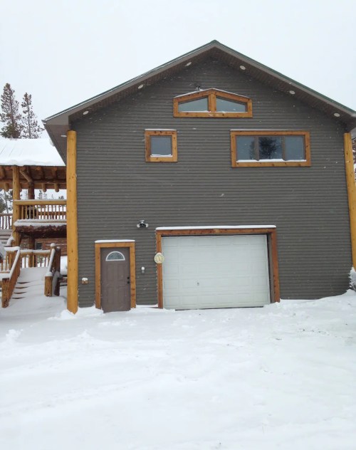 A two-story gray house with wooden accents, surrounded by snow, featuring a garage and a small front door.