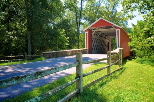 A red covered bridge spans a road, surrounded by lush greenery and a wooden fence.