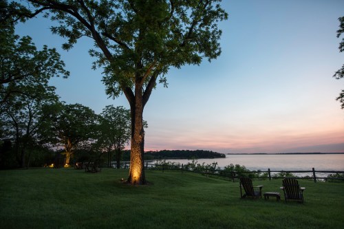 A serene lakeside view at dusk, featuring trees, green grass, and two wooden chairs facing the water.