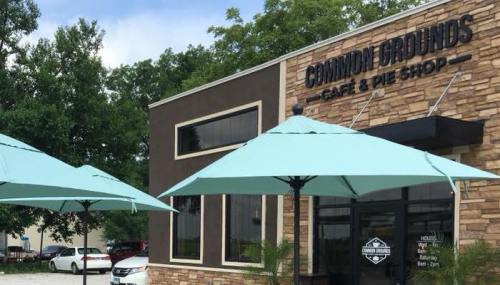 Exterior of Common Grounds Café & Pie Shop, featuring blue umbrellas and a stone facade. Trees in the background.