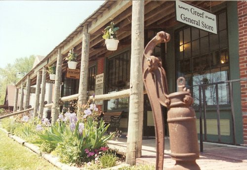 A rustic general store with wooden pillars, flower pots, and a vintage hand pump in the foreground.