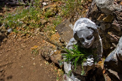 A weathered stone statue of a girl holding a bouquet of wildflowers, surrounded by a natural, earthy landscape.