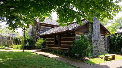 A rustic log cabin surrounded by greenery, featuring a stone chimney and a pathway leading to the entrance.