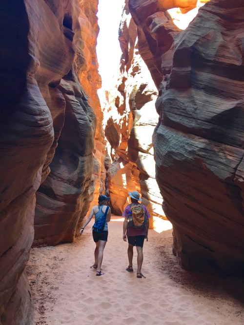 Two hikers walk through a narrow, sunlit canyon with towering rock formations on either side.