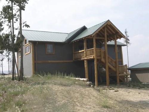 A modern cabin with a wooden porch and green roof, set in a natural landscape with sparse vegetation.