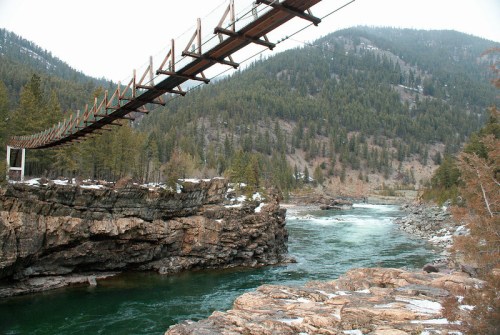 A suspension bridge spans a river, surrounded by rocky cliffs and lush green mountains under a cloudy sky.