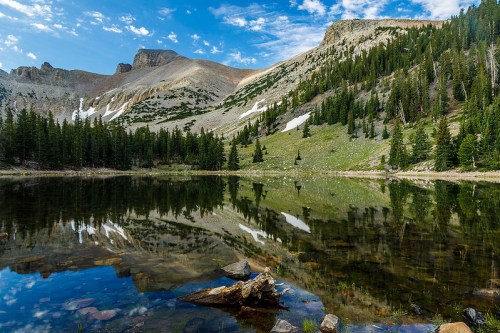A serene mountain landscape with a clear lake reflecting the surrounding trees and rocky peaks under a blue sky.