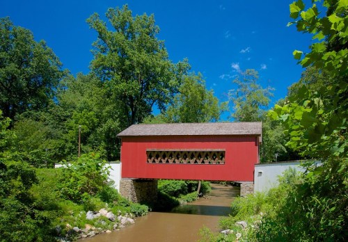 A red covered bridge spans a creek, surrounded by lush green trees and a clear blue sky.