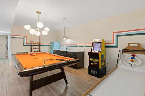 A game room featuring a pool table, arcade machine, and retro decor with colorful wall stripes.