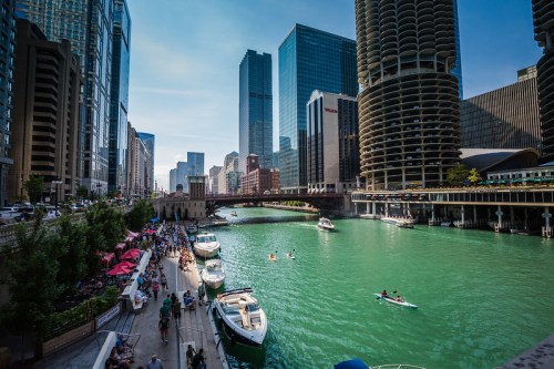 A vibrant cityscape of Chicago with boats on a green river, surrounded by tall buildings and people enjoying the waterfront.