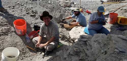 Three people excavating at a dig site, surrounded by tools and buckets, focused on their work in a rocky area.