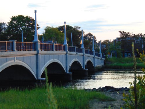 A stone bridge arches over a calm waterway, surrounded by greenery and trees under a soft evening sky.
