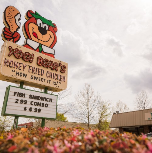 Sign for Yogi Bear's Honey Fried Chicken featuring a cartoon bear and menu items, surrounded by greenery and cloudy sky.