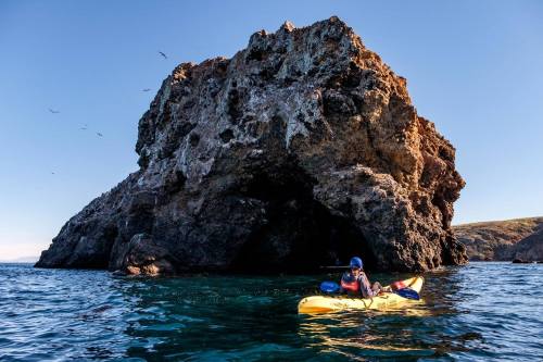 A person in a kayak paddles near a large rocky outcrop in clear blue water under a sunny sky.
