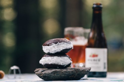 Two chocolate whoopie pies with cream filling on a wooden surface, with a beer bottle and glass in the background.