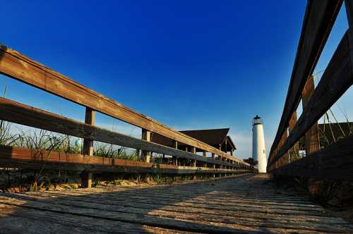 A wooden walkway leads to a lighthouse under a clear blue sky, surrounded by grass and sandy terrain.