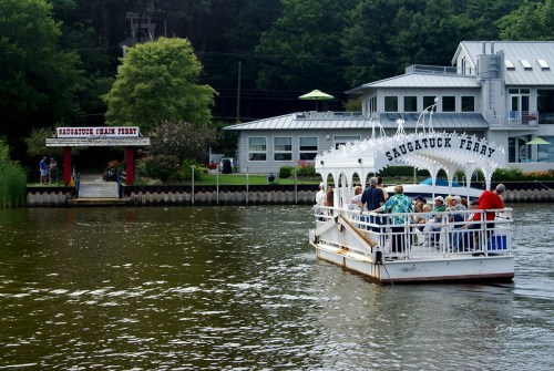 A ferry with passengers approaches a dock labeled "Saugatuck Chain Ferry" on a calm river surrounded by greenery.