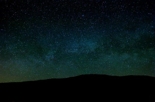 A starry night sky filled with stars and a faint Milky Way, silhouetted by a dark mountain range below.