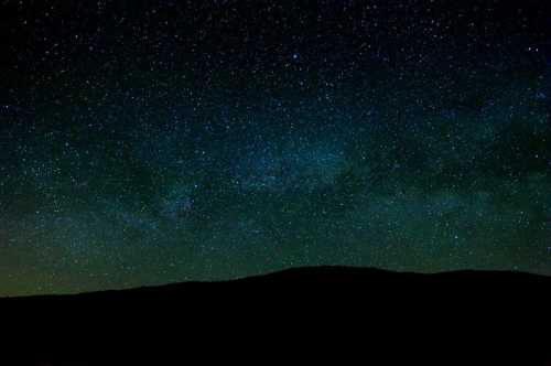 A starry night sky filled with stars and a faint Milky Way, silhouetted by a dark mountain range below.