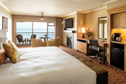 Cozy hotel room with a king bed, seating area, desk, and ocean view through large windows.