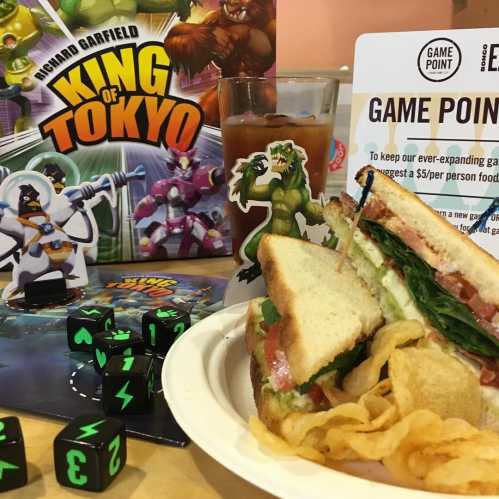 A plate of sandwiches and chips beside a board game, with a drink and dice on a table featuring a colorful game backdrop.