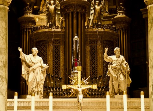 Two marble statues of saints stand on either side of a cross, with ornate architecture and candlelight in the background.