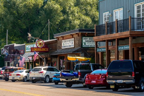 A row of rustic shops with American flags, parked cars, and a kayak on top, set in a small town.