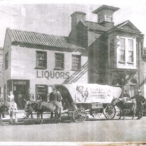 Historic black-and-white photo of a liquor store with a horse-drawn wagon and people standing outside.