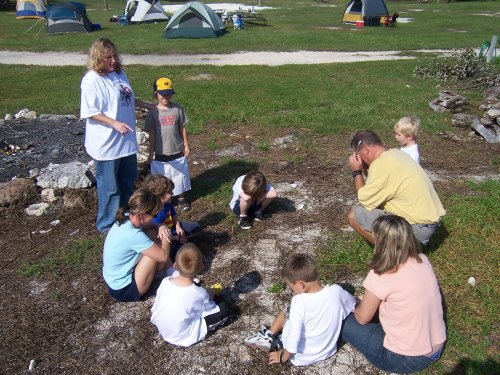 A group of children and adults gather around a campfire site, engaging in an outdoor activity at a campsite.
