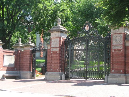 Ornate wrought iron gates with brick pillars, surrounded by lush greenery, leading to a historic campus entrance.