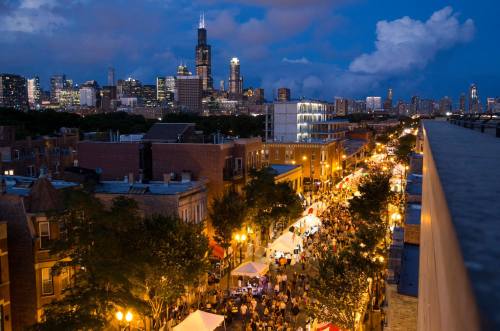 A vibrant street festival at night, with crowds and tents, set against a skyline of Chicago's tall buildings.