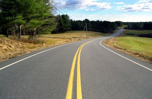 A winding road stretches through a rural landscape, flanked by trees and open fields under a blue sky.