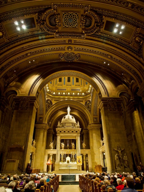 Interior of a grand church with ornate architecture, featuring a large altar and a congregation seated in pews.