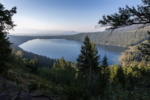 A serene lake surrounded by lush forests and mountains under a clear blue sky at dawn.