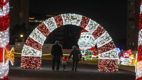 A family walks under a festive candy cane arch adorned with colorful lights during a holiday display.