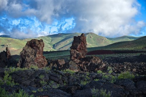 Lava rock formations stand against a backdrop of green hills and a blue sky with scattered clouds.