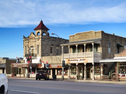 Historic buildings line a street, featuring shops with signs, a tower, and a clear blue sky above.
