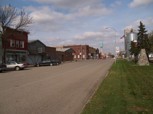 A quiet street lined with small buildings and parked cars, under a cloudy sky. A water tower is visible in the distance.