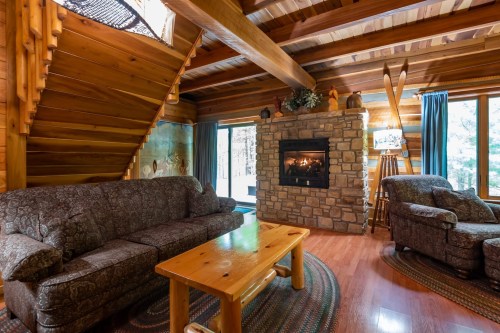 Cozy log cabin living room with a stone fireplace, wooden beams, and comfortable furniture. Natural light from large windows.