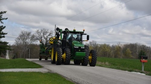 A green tractor drives down a rural road, with fields and trees in the background under a cloudy sky.